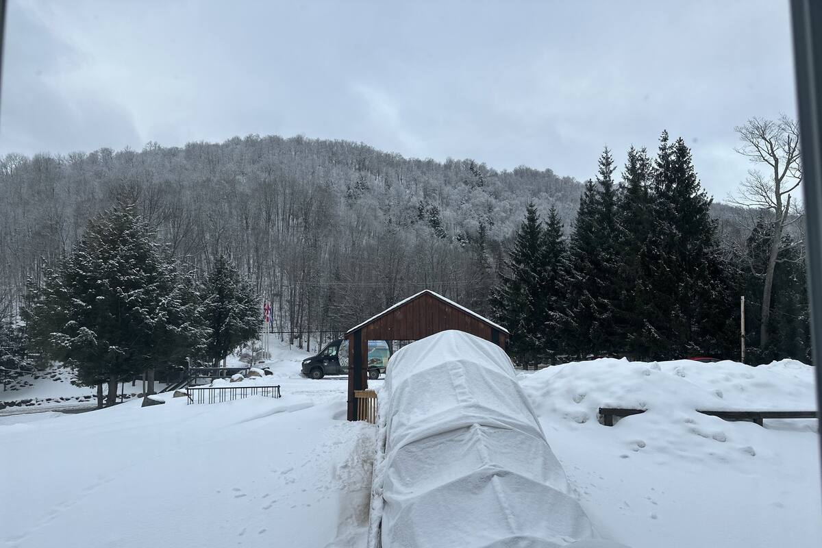 Schneebedeckte Landschaft mit einer Holzhütte und einem überdachten Fahrzeug im Vordergrund.