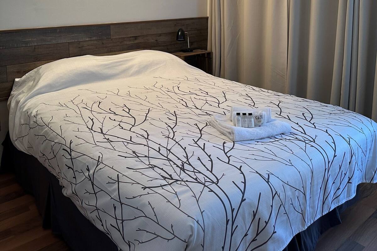 Ein ordentlich bezogenes Bett mit Bettdecke mit Baumzweigmuster, ein Nachttisch mit Lampe und ein Fenster mit beigen Vorhängen.