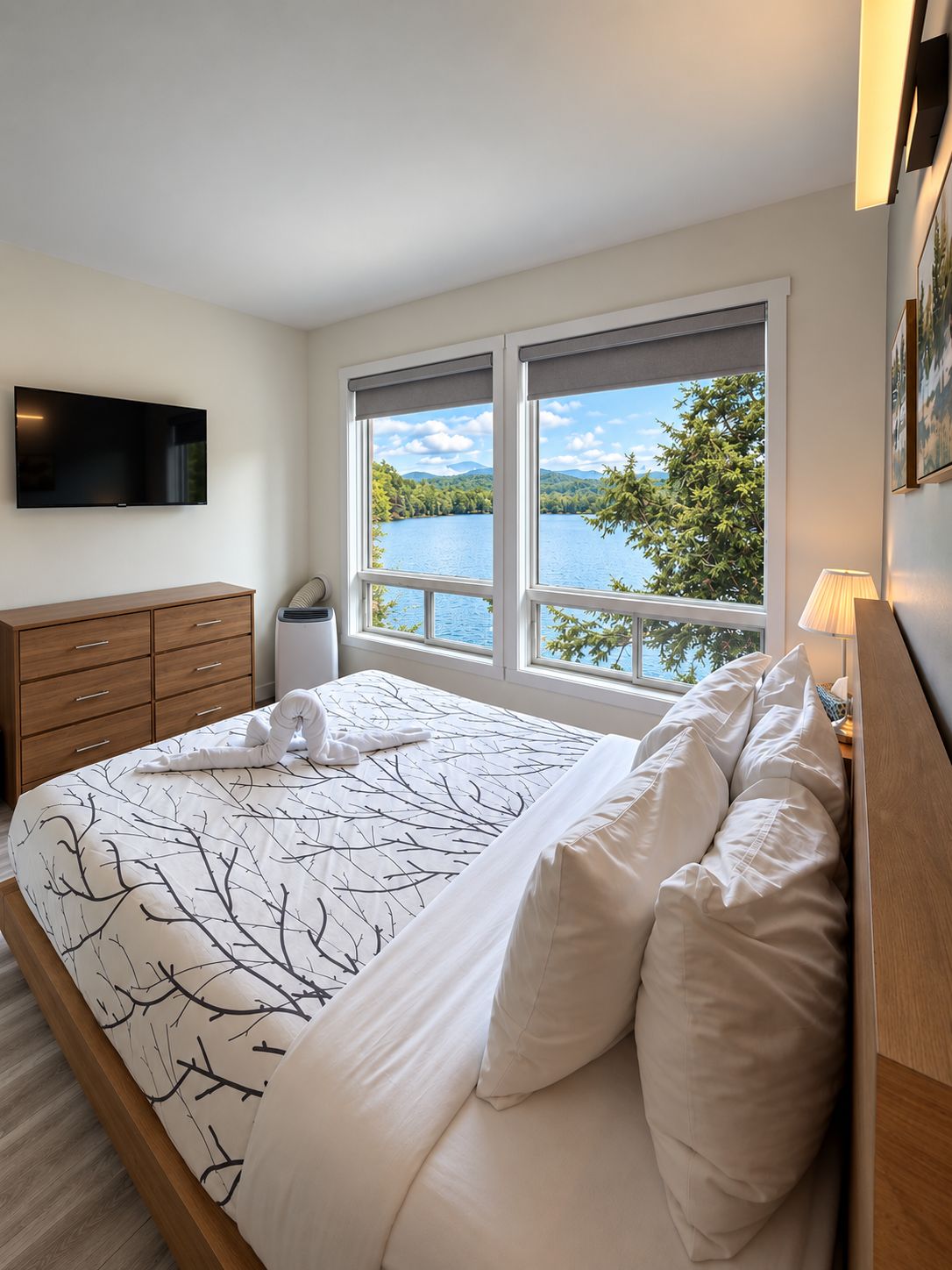A bedroom with a bed, a TV, a window with a view of a lake, and a wooden dresser.