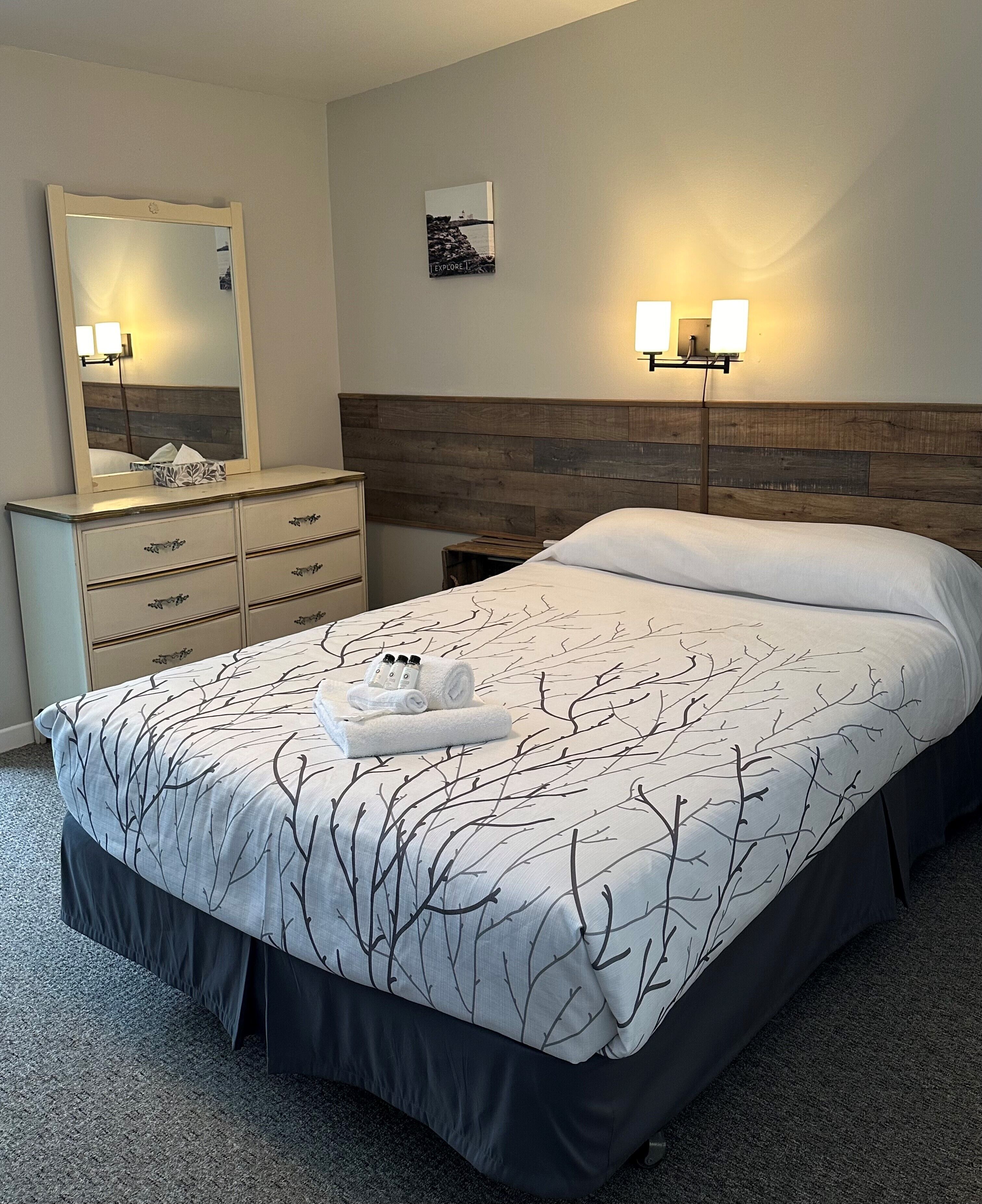 A neatly made bed with a tree branch pattern bedspread, a wooden headboard, a bedside table with a lamp, and a dresser with drawers.