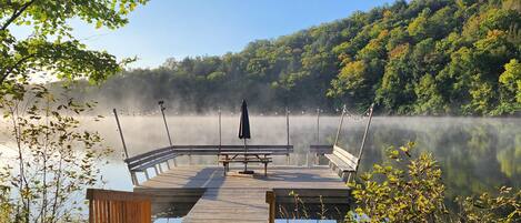 A wooden dock extending into a calm lake with a forested hillside in the background.