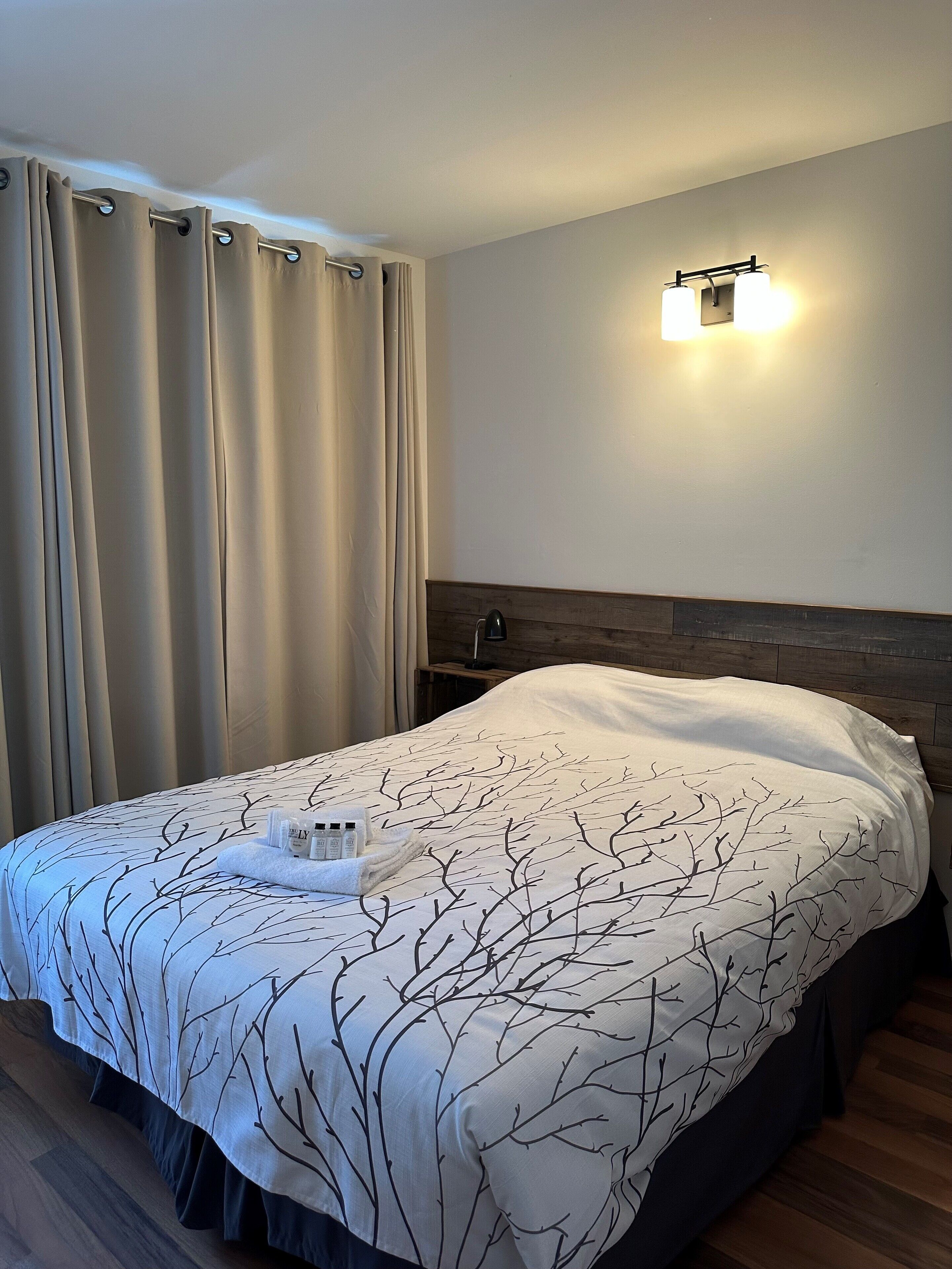 Un lit bien fait, recouvert d’une couvre-lit à motifs de branches d’arbres, une table de chevet et un luminaire fixé au mur.
