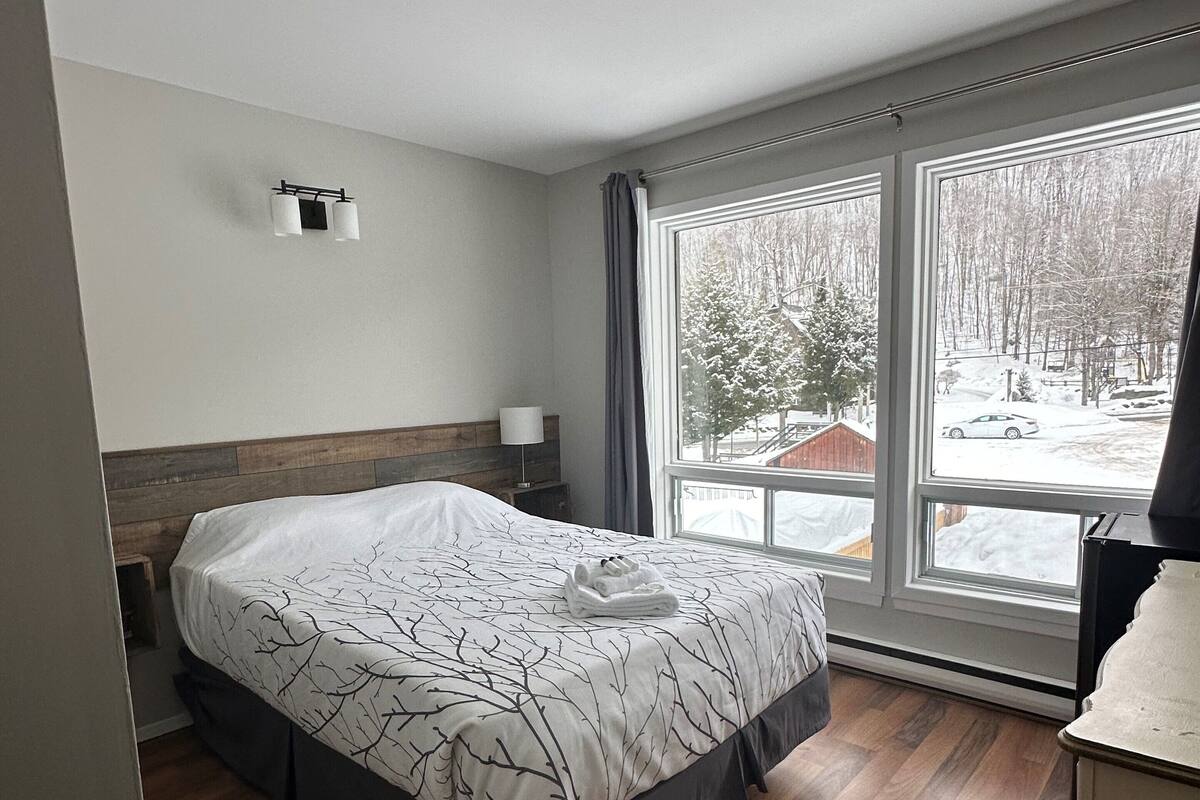 Ein modernes Schlafzimmer mit einem großen Bett, einem Holzkopfende, einem Nachttisch mit Lampe und Blick auf eine verschneite Landschaft durch große Fenster.