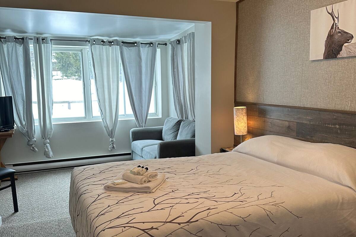 Ein Hotelzimmer mit einem großen Bett, einer Sitzecke inklusive Sessel und Tisch, einem Fenster mit Vorhängen sowie einem an der Wand befestigten Bild von einem Reh.