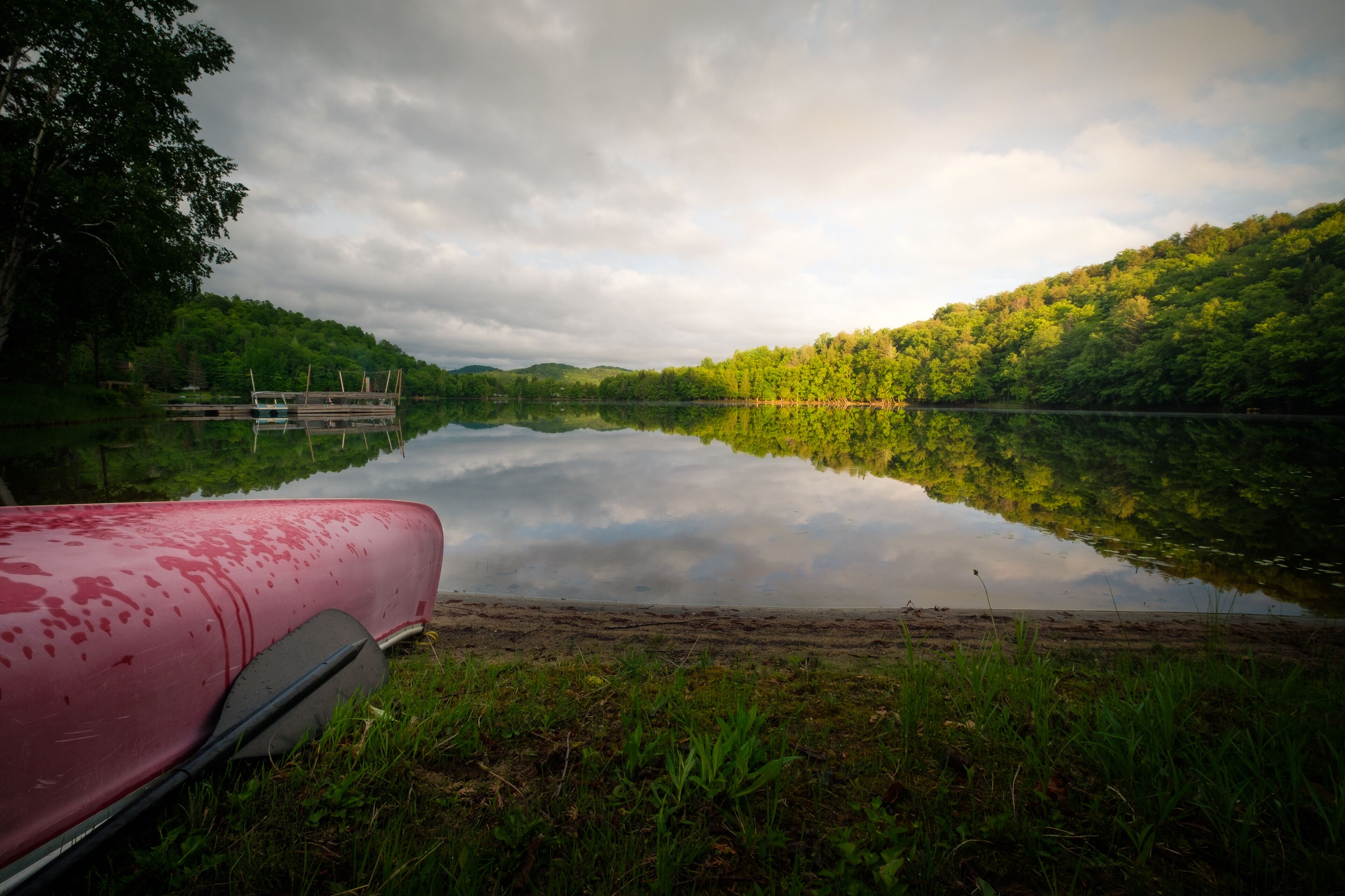 A calm lake with a red canoe on the shore, surrounded by lush greenery and a cloudy sky.