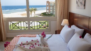 A hotel room with a bed, a window with a view of the beach and ocean, and a nightstand with a lamp. - Oyster Bay Beach Suites (Marsa Alam)