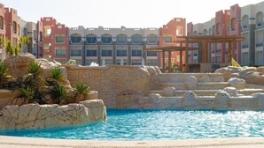 A pool area with rock formations and palm trees, surrounded by multi-story buildings with balconies. - Oyster Bay Beach Suites (Marsa Alam)