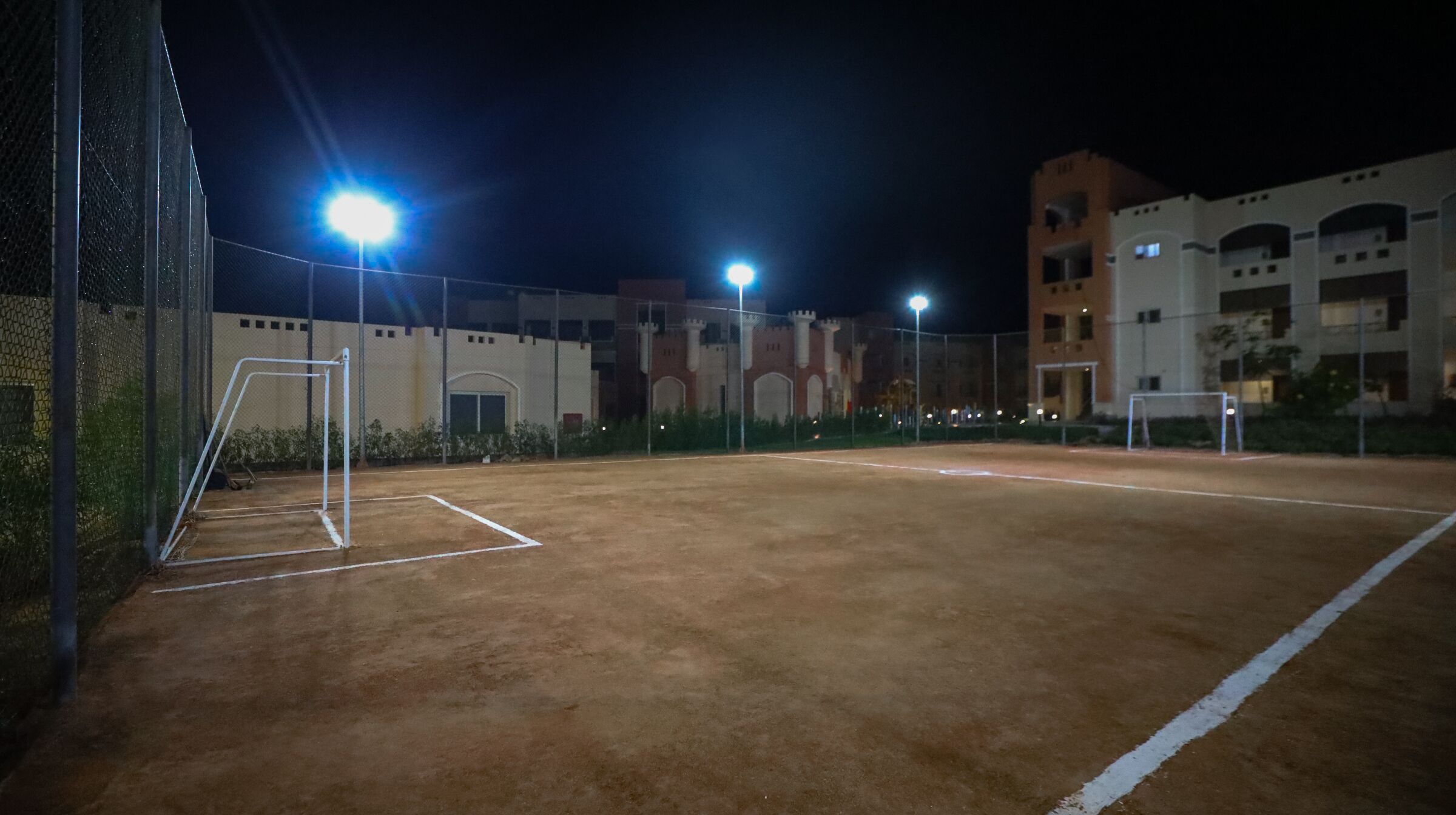An outdoor tennis court at night with floodlights, surrounded by a fence and buildings in the background.