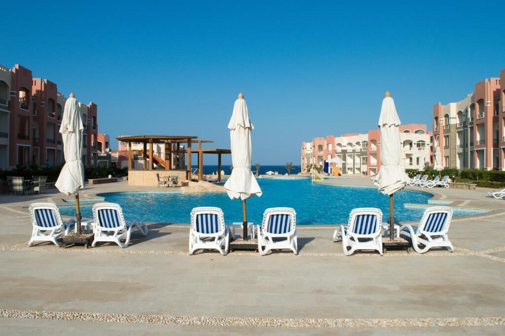 Uma área à beira da piscina com espreguiçadeiras e guarda-sóis, uma piscina e edifícios ao fundo.