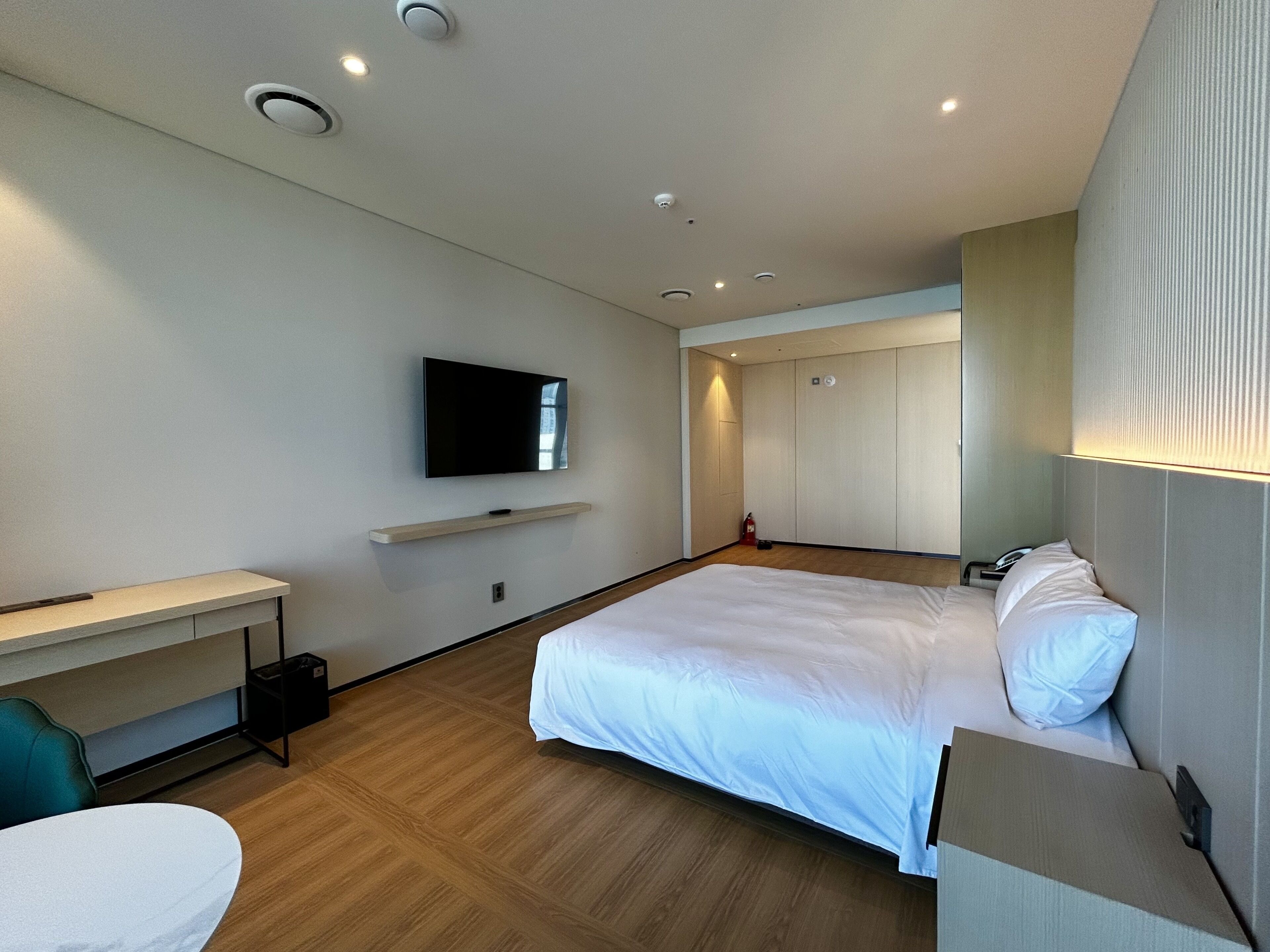 A modern hotel room with a large bed, a flat-screen TV mounted on the wall, a bedside table, and a wooden floor.