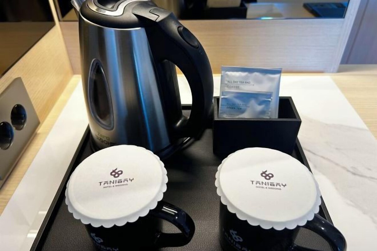 A kitchen counter with a stainless steel kettle, two black mugs with white lids, and a black tray with a tea bag and a card.