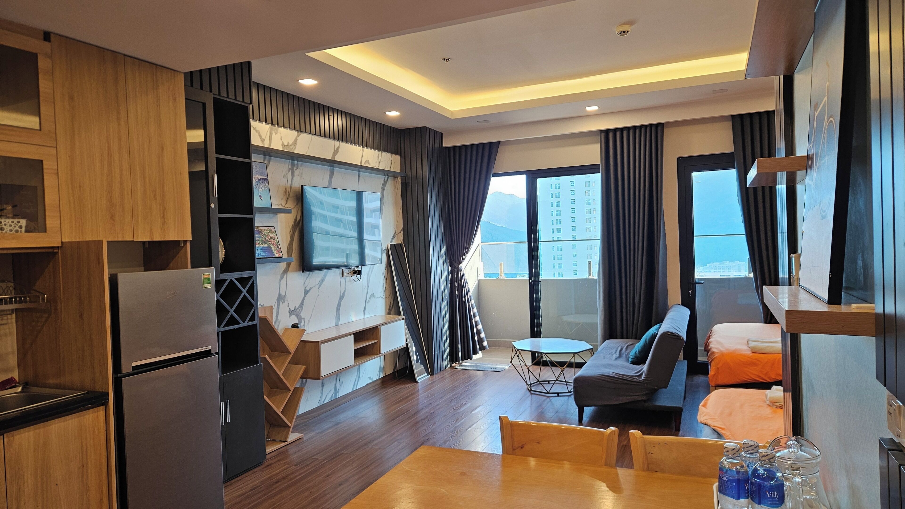 Una habitación de hotel moderna con cocina, zona de estar con sofá y mesa de comedor, televisor montado en la pared y balcón con vistas a la ciudad.