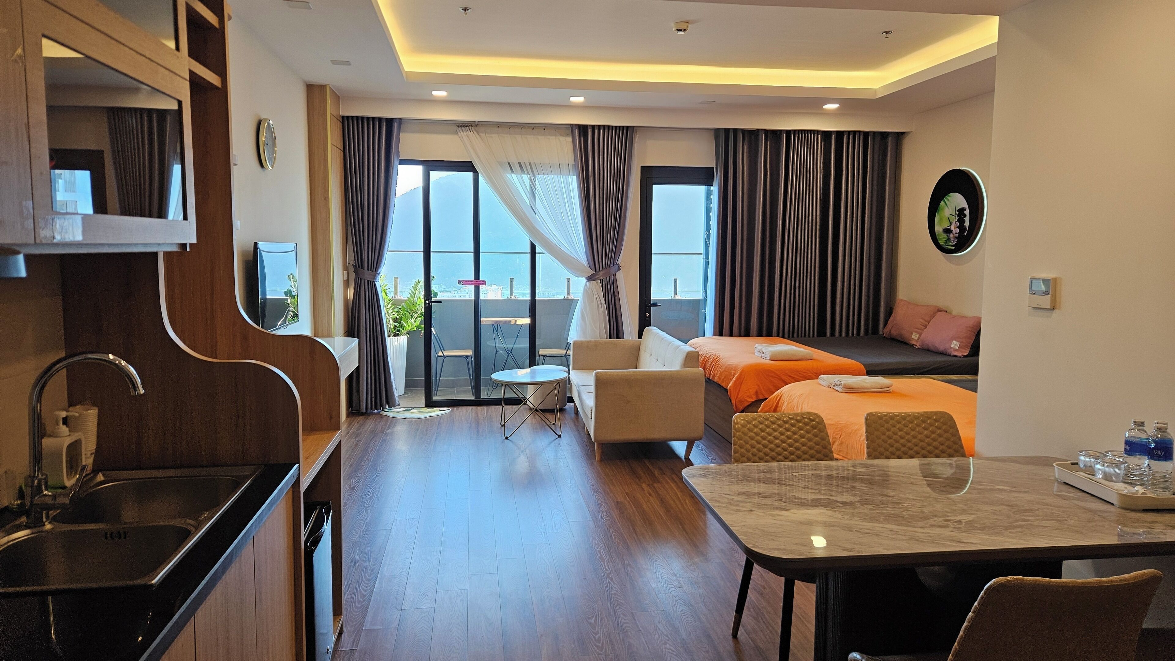 Une chambre d’hôtel avec un plancher en bois, une table à manger, un canapé, un lit et un balcon avec des rideaux.