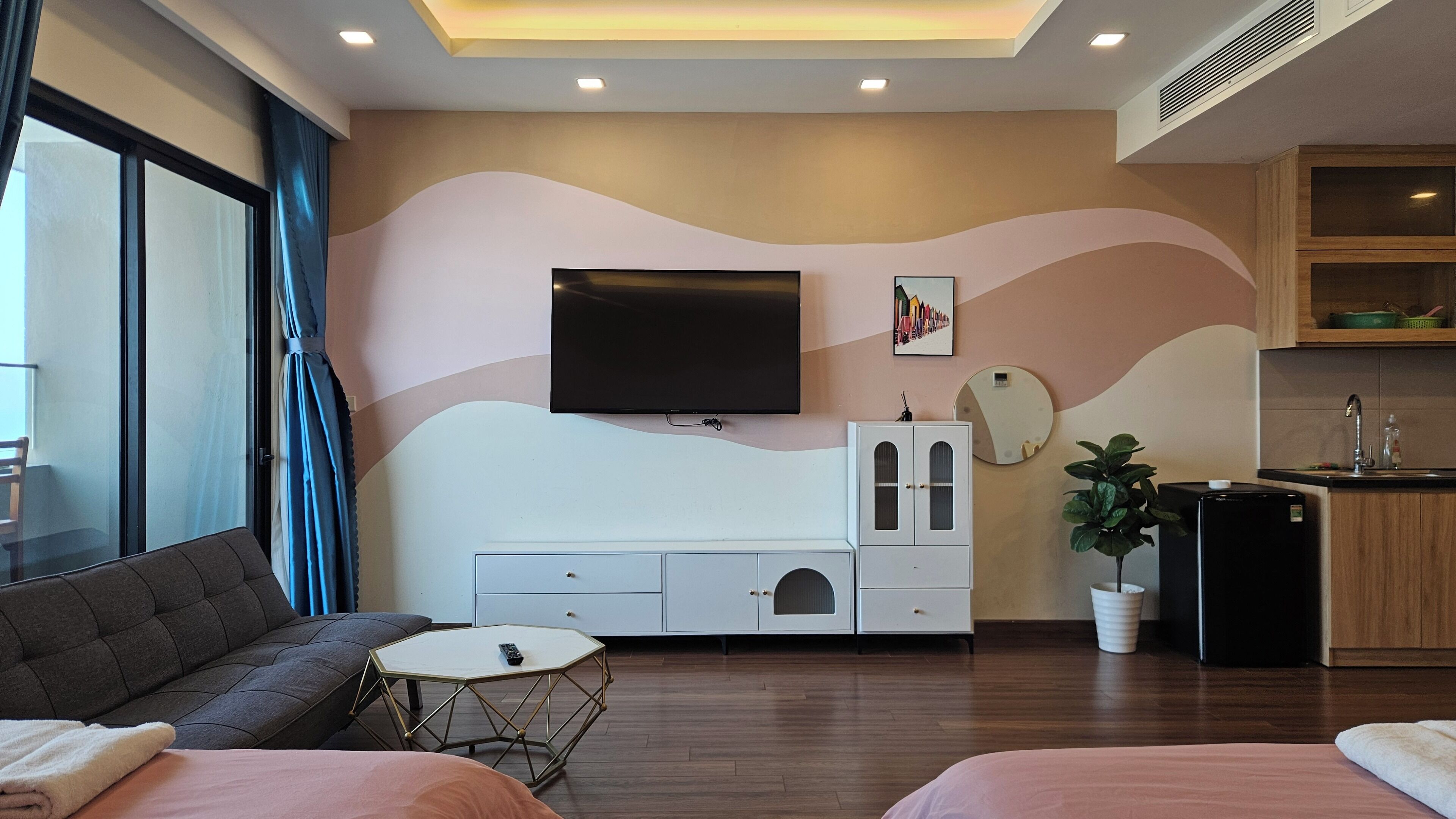 A modern hotel room with a sofa, a small round table, a TV mounted on the wall, a cabinet with drawers, a potted plant, and a kitchen area with a sink and a refrigerator.