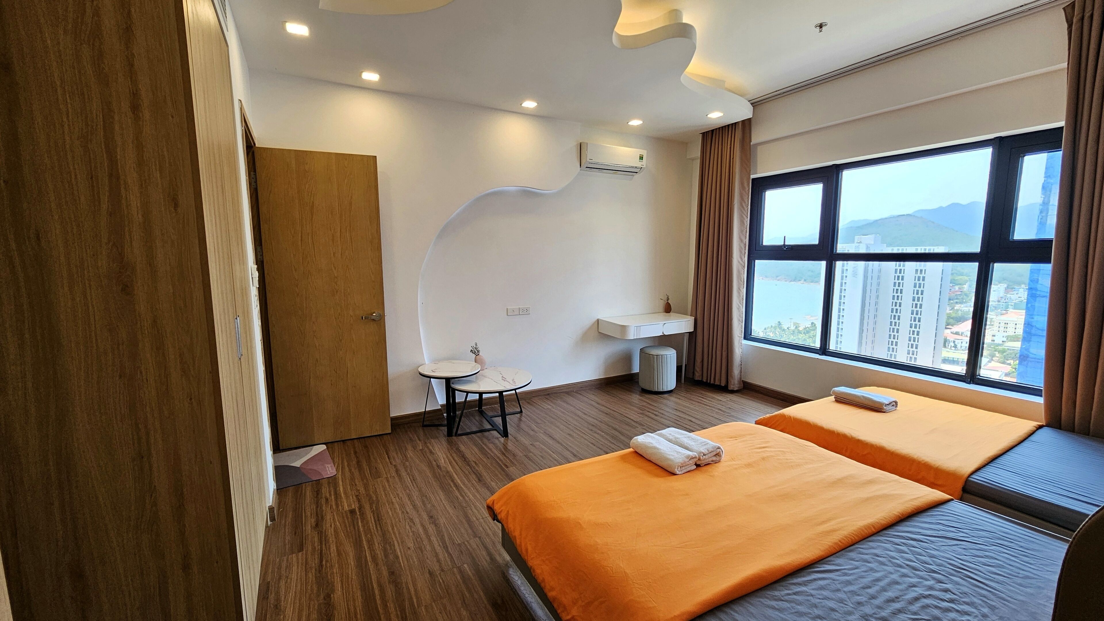 A hotel room with two beds, a small table, a sink, and a view of the city.