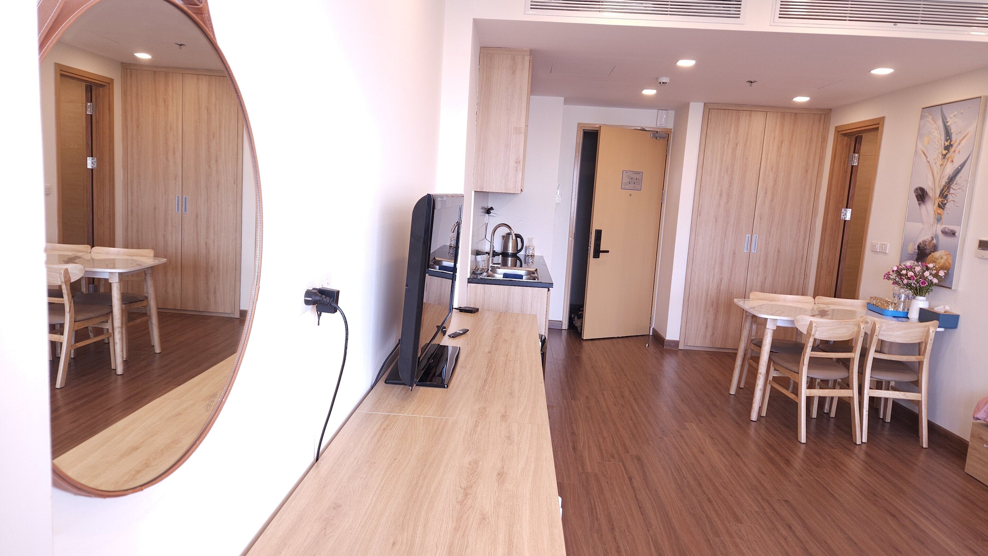 A modern room with wooden flooring, a dining table with chairs, a kitchen area, and a large mirror.