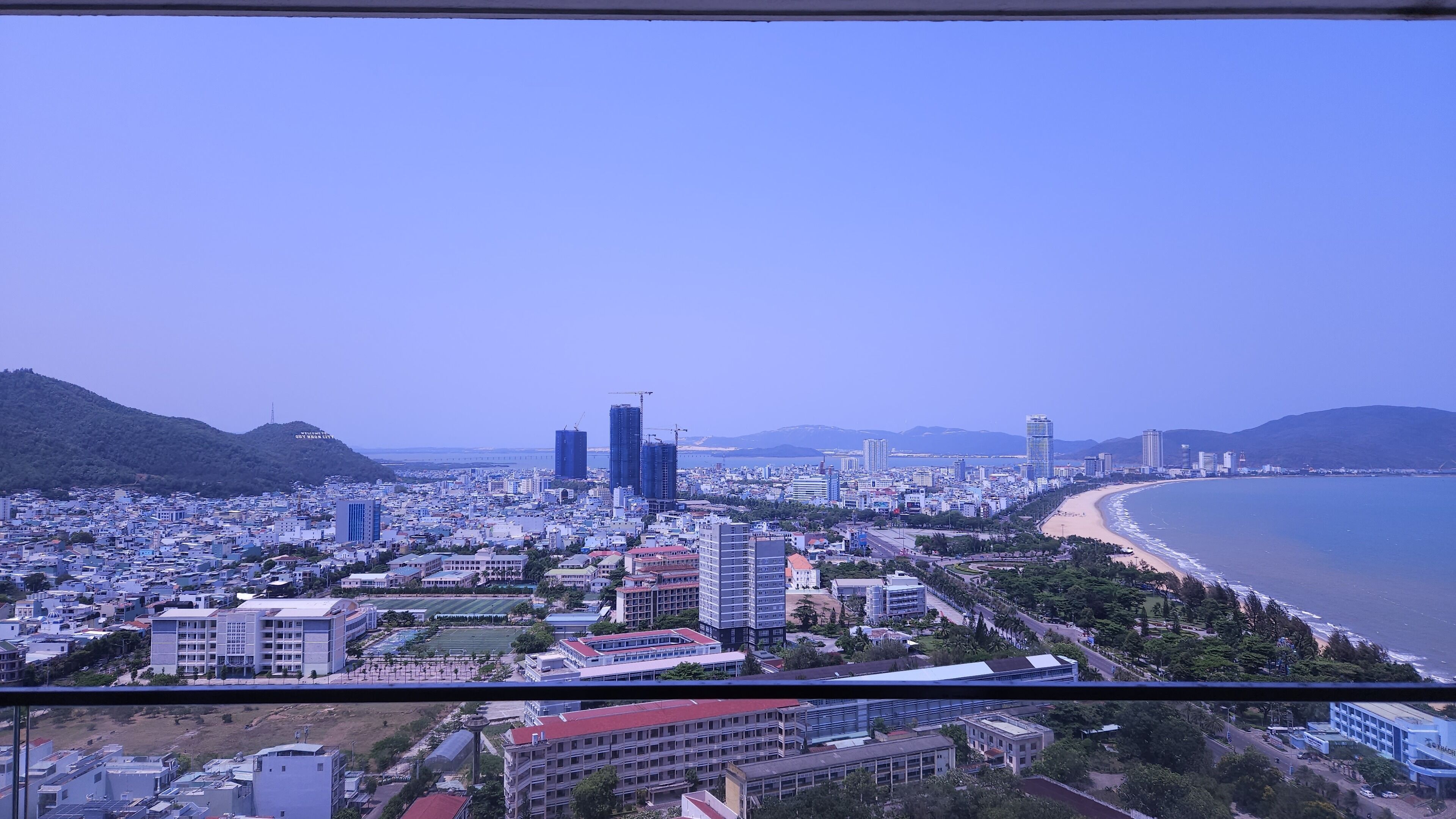 A coastal cityscape with a mix of modern and traditional buildings, a beach, and mountains in the background.