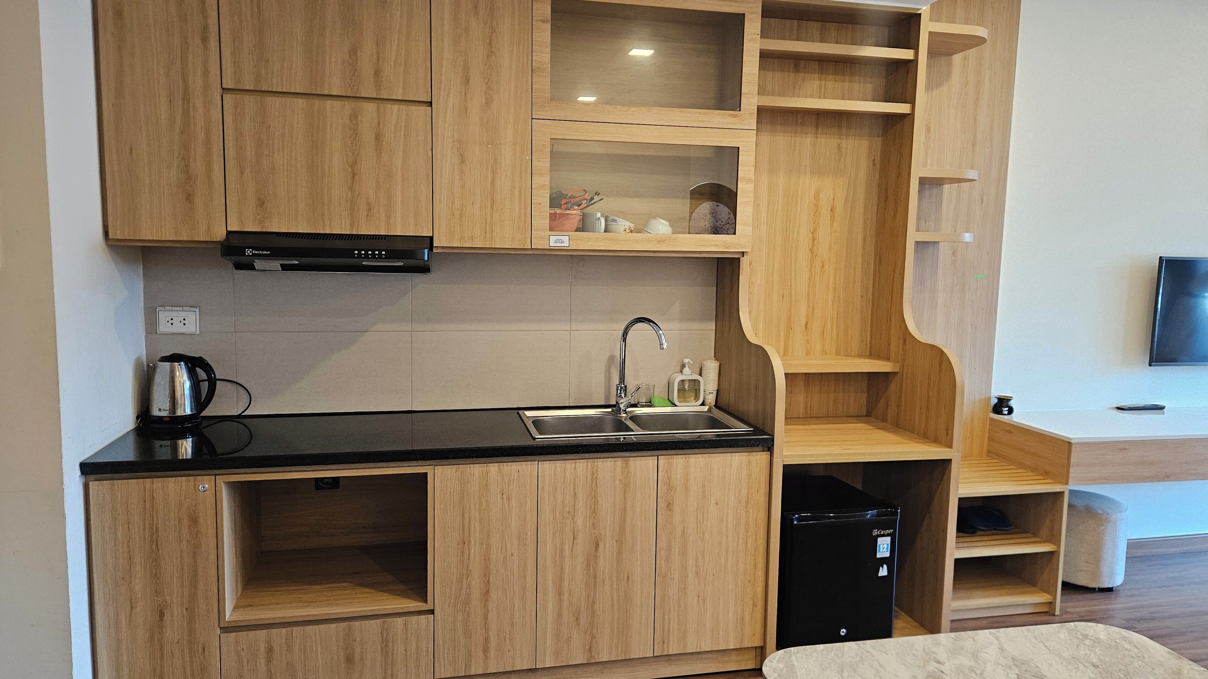 A modern kitchen with wooden cabinets, a black countertop, a built-in dishwasher, and a flat-screen TV mounted on the wall.