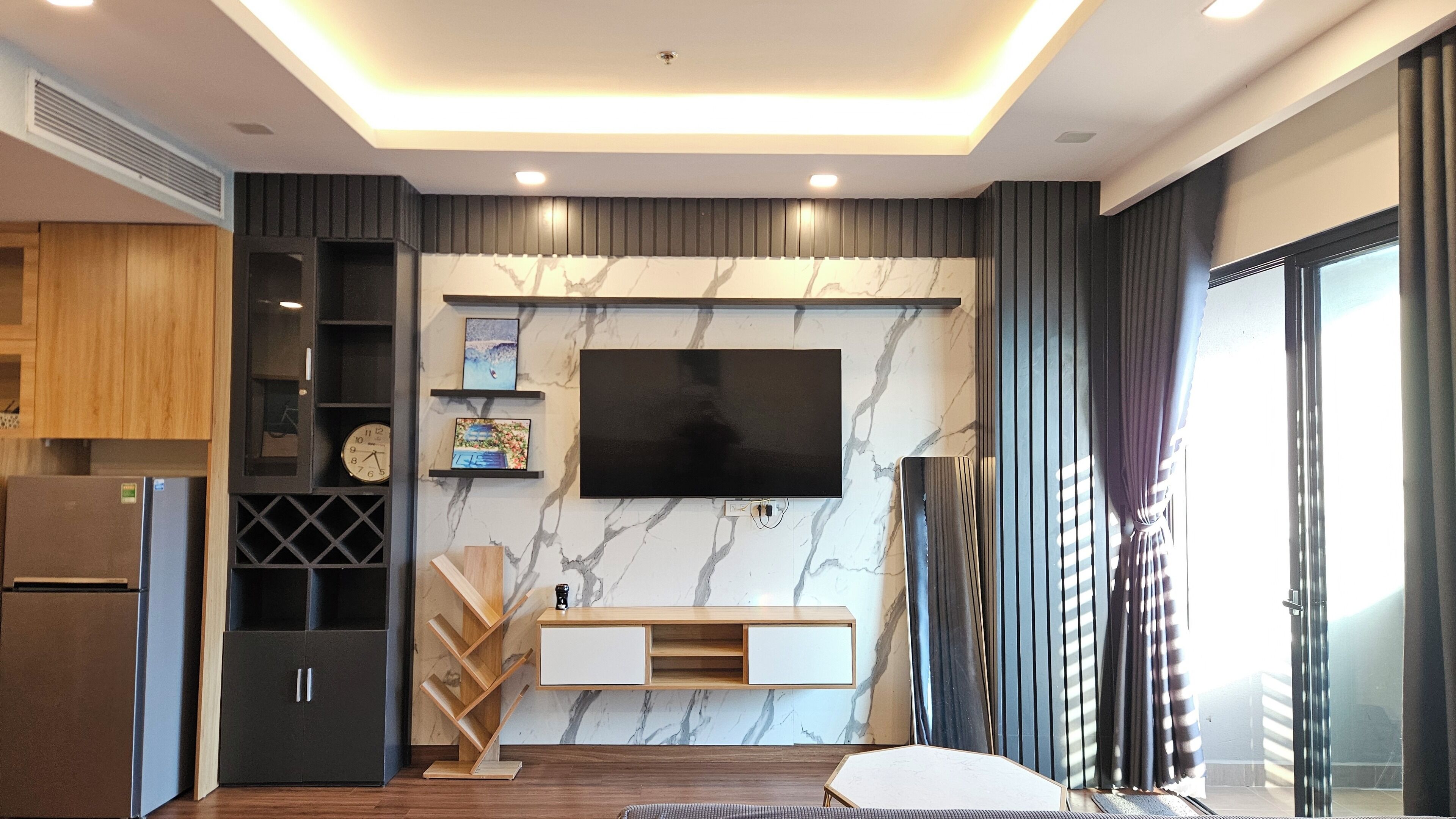Un salon moderne doté d’un téléviseur à écran plat fixé sur un mur en marbre, d’étagères en bois, d’une machine à café intégrée et d’un coin salon avec un canapé et une table basse.