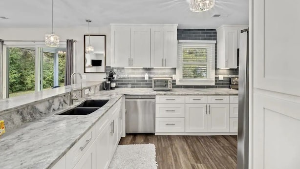 A modern kitchen with white cabinets, a marble countertop, stainless steel appliances, and a large window offering a view of greenery.