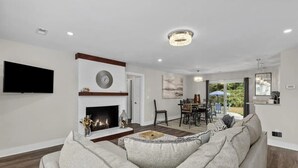 A modern living room with a fireplace, a flat-screen TV, and a dining area visible through an open door.