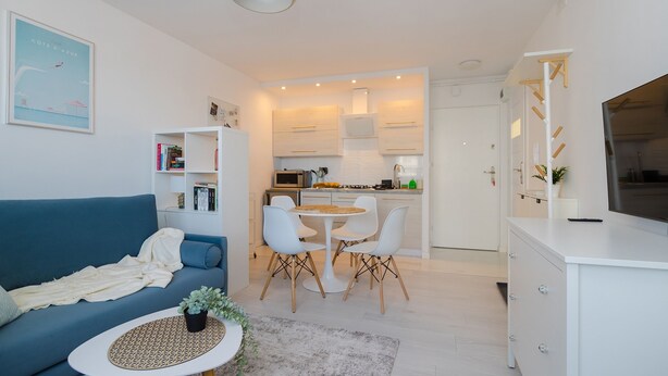 A modern apartment with a blue sofa, a dining table with chairs, a kitchen area, and a TV on a cabinet. - ShortStayPoland Batorego B139 (Warsaw)