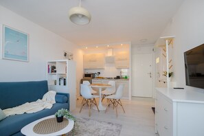 A modern apartment with a blue sofa, a dining table with chairs, a kitchen area, and a TV on a cabinet.