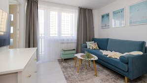 A modern living room with a blue sofa, a small table with a plant, a white TV stand, and a window with sheer curtains. - ShortStayPoland Batorego B139 (Warsaw)