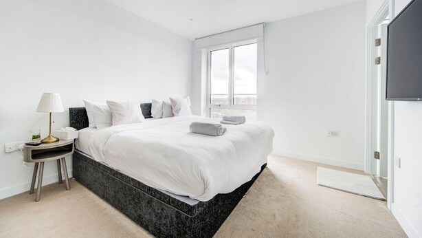A modern hotel room with a large bed, a bedside table with a lamp, a flat-screen TV, and a window with a city view.