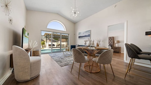 A spacious living room with a large round table, comfortable seating, a view of a pool, and a modern chandelier. - 8615 Vacation Oasis House Private Pool Grill (Kissimmee)