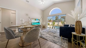 A modern living room with a dining area, a large window with a view of palm trees, and a ceiling light fixture. - 8615 Vacation Oasis House Private Pool Grill (Kissimmee)