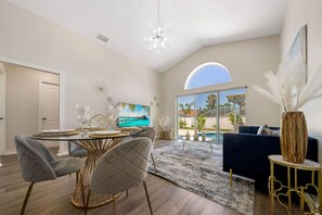 Una sala de estar moderna con comedor, un ventanal con vista a palmeras y un lámpara en el techo.