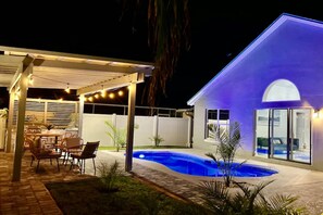 Un patio trasero con piscina, un área de patio cubierta con luces colgantes y una casa con puerta de vidrio.