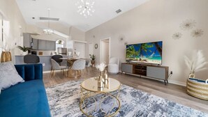 A spacious living room with a blue sofa, a coffee table, and a large flat-screen TV displaying a beach scene. - 8615 Vacation Oasis House Private Pool Grill (Kissimmee)