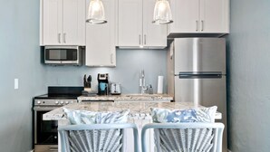 Una cocina con armarios blancos, un refrigerador de acero inoxidable, un microondas, una estufa y dos sillas de mimbre con una mesa.