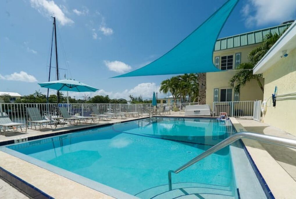 A clear blue swimming pool with lounge chairs and umbrellas, surrounded by a white fence and palm trees.