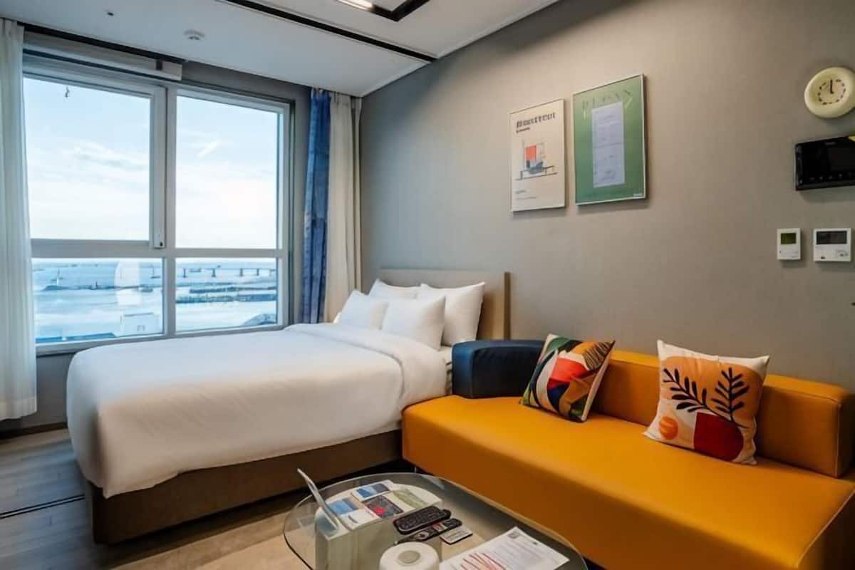 Habitación de hotel con cama, sofá, ventana con vistas, un reloj de pared y una pequeña mesa con objetos encima.