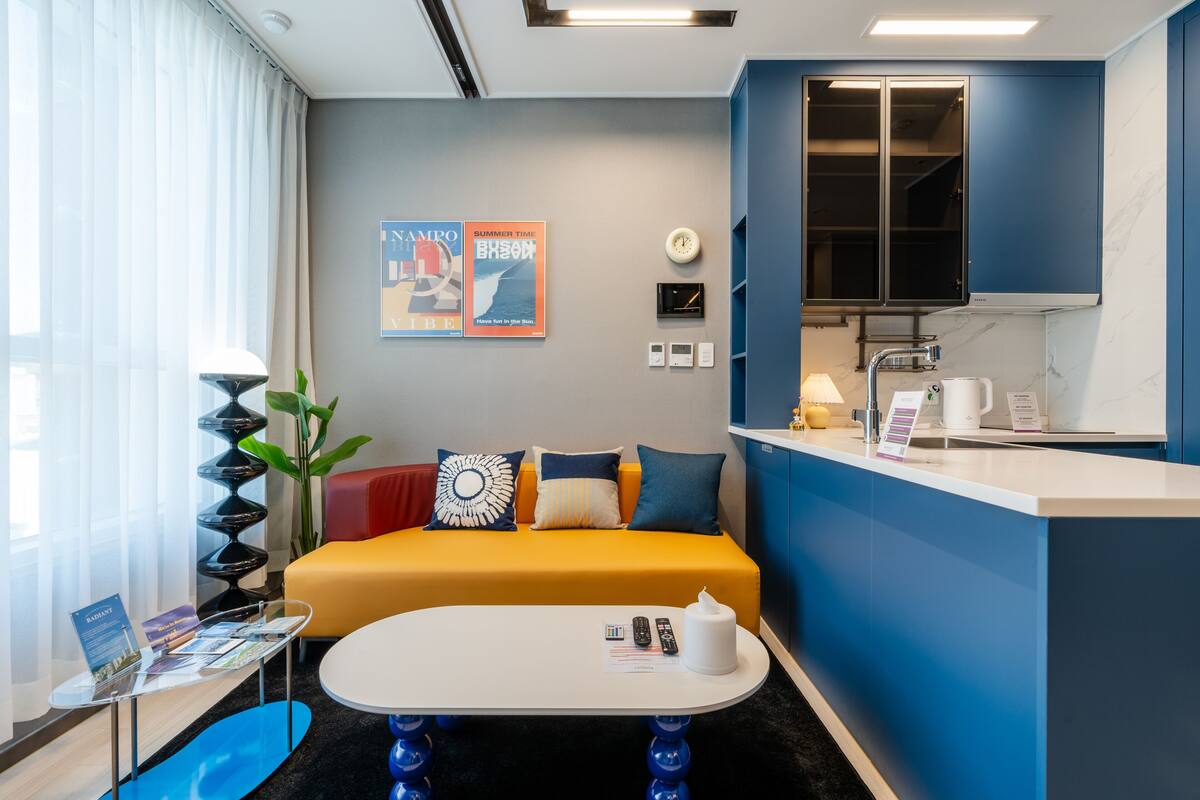 Una habitación de hotel moderna con un sofá amarillo, una pequeña mesa redonda, una zona de cocina con fregadero y encimera, y un gran ventanal con cortinas translúcidas.