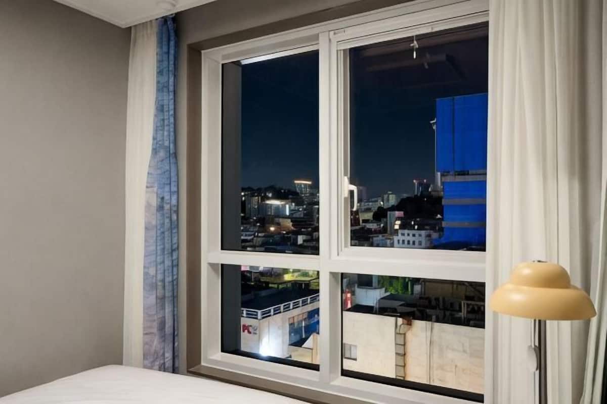 Un dormitorio con vista a la ciudad a través de una ventana, una cama y una lámpara.