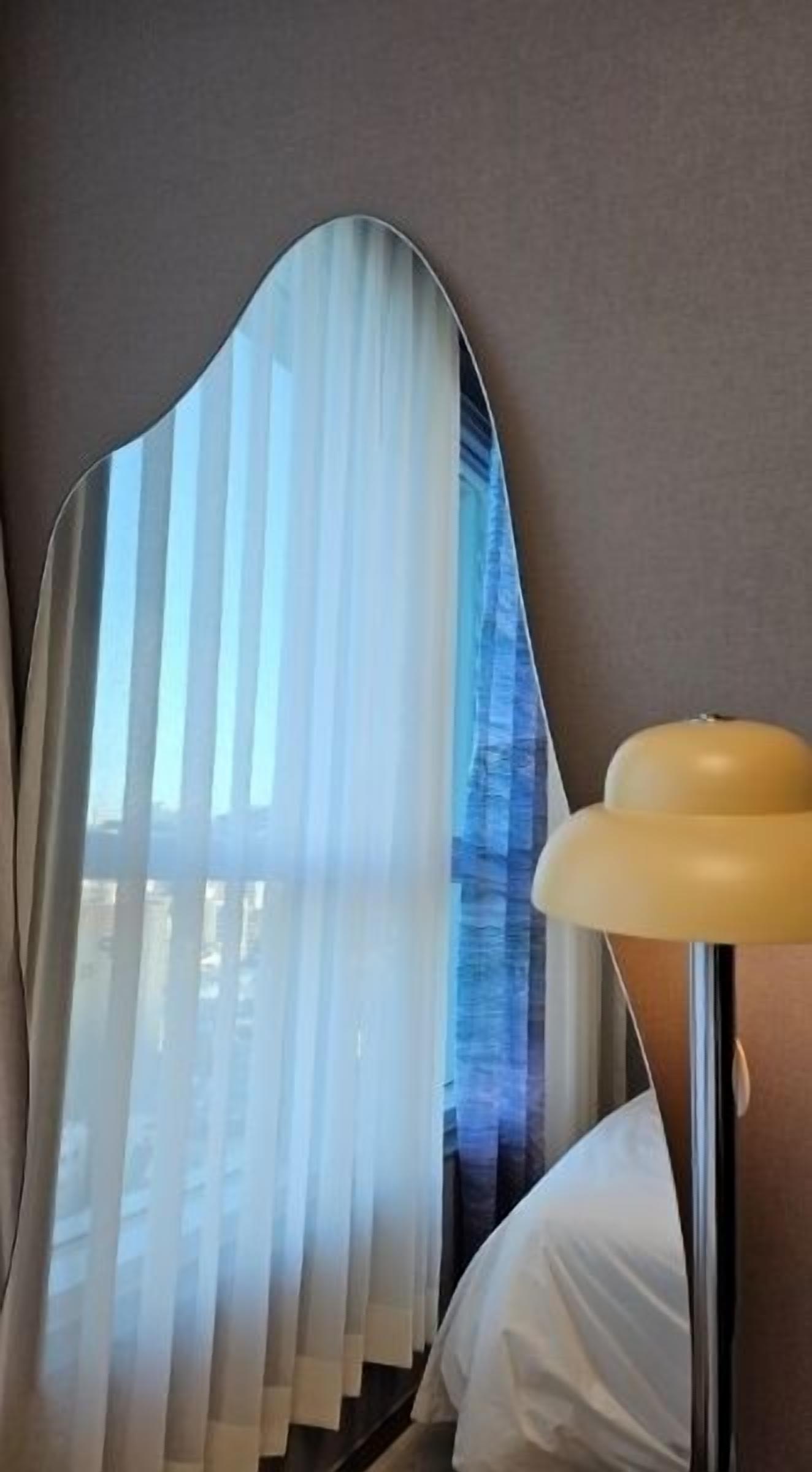 A room with a large mirror reflecting a window with sheer curtains, a bedside lamp, and a white bed.