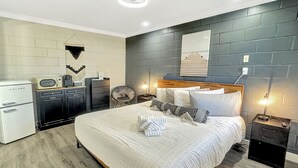 A modern bedroom with a large bed, a small kitchenette, a chair, and a nightstand with a lamp. - King Room Hotel- 2 Pools, Near Beach/Boardwalk, No Pets Allowed (Carolina Beach)