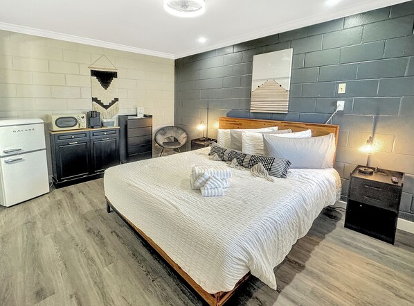 A modern bedroom with a large bed, a small kitchenette, a chair, and a nightstand with a lamp.