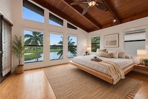 A bedroom with a large bed, multiple windows, a ceiling fan, and a view of palm trees.