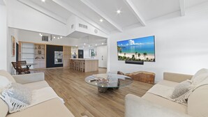 Ein geräumiger Wohnbereich mit einem Glastisch, beigefarbenen Sofas und einem großen Fernseher, der eine Strandansicht zeigt.