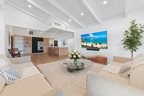 A spacious living room with a glass coffee table, beige sofas, a dining area, and a kitchen in the background.
