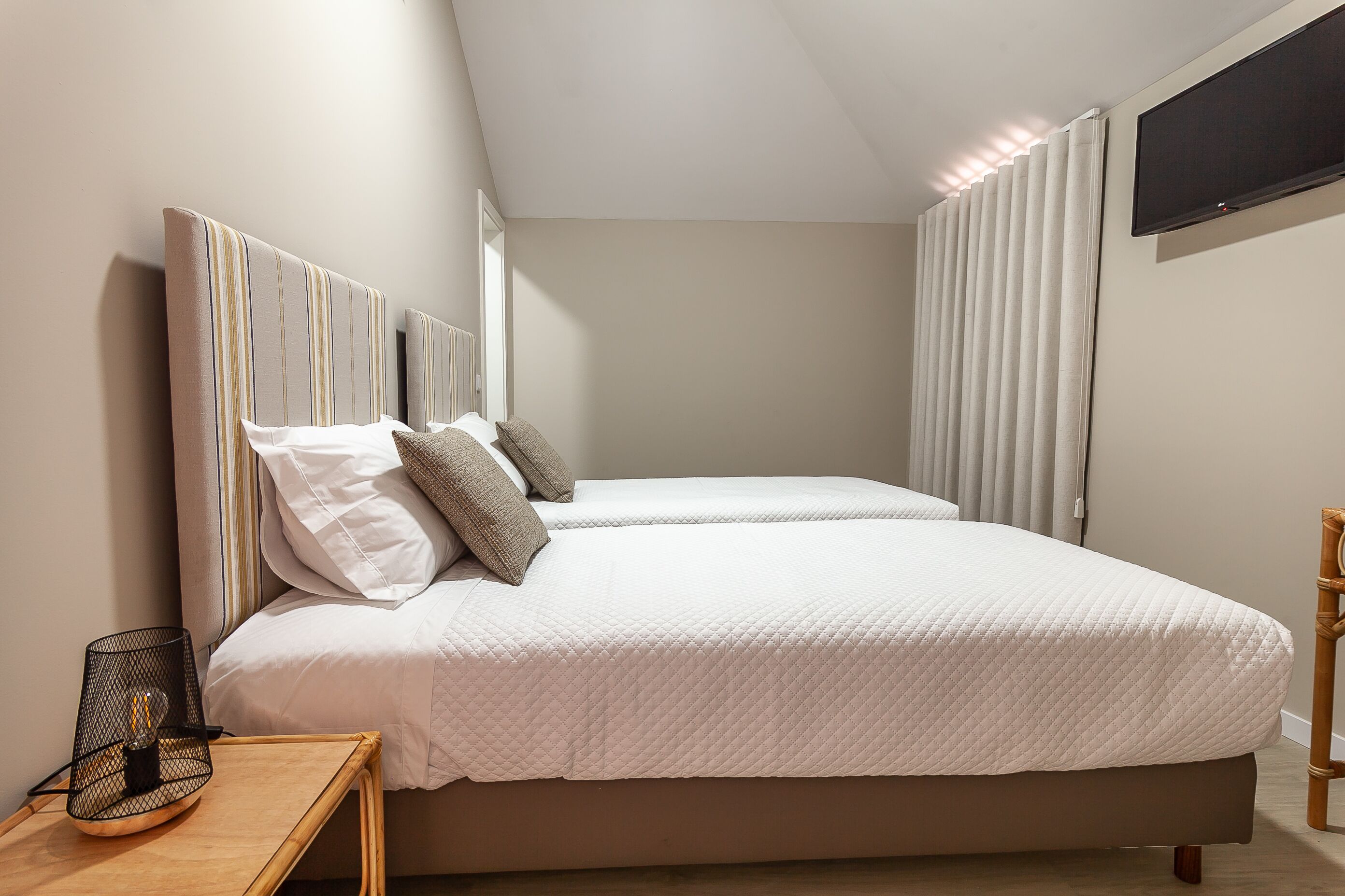 Cama doble con edredón blanco acolchado, mesita de noche con lámpara y televisor montado en la pared.