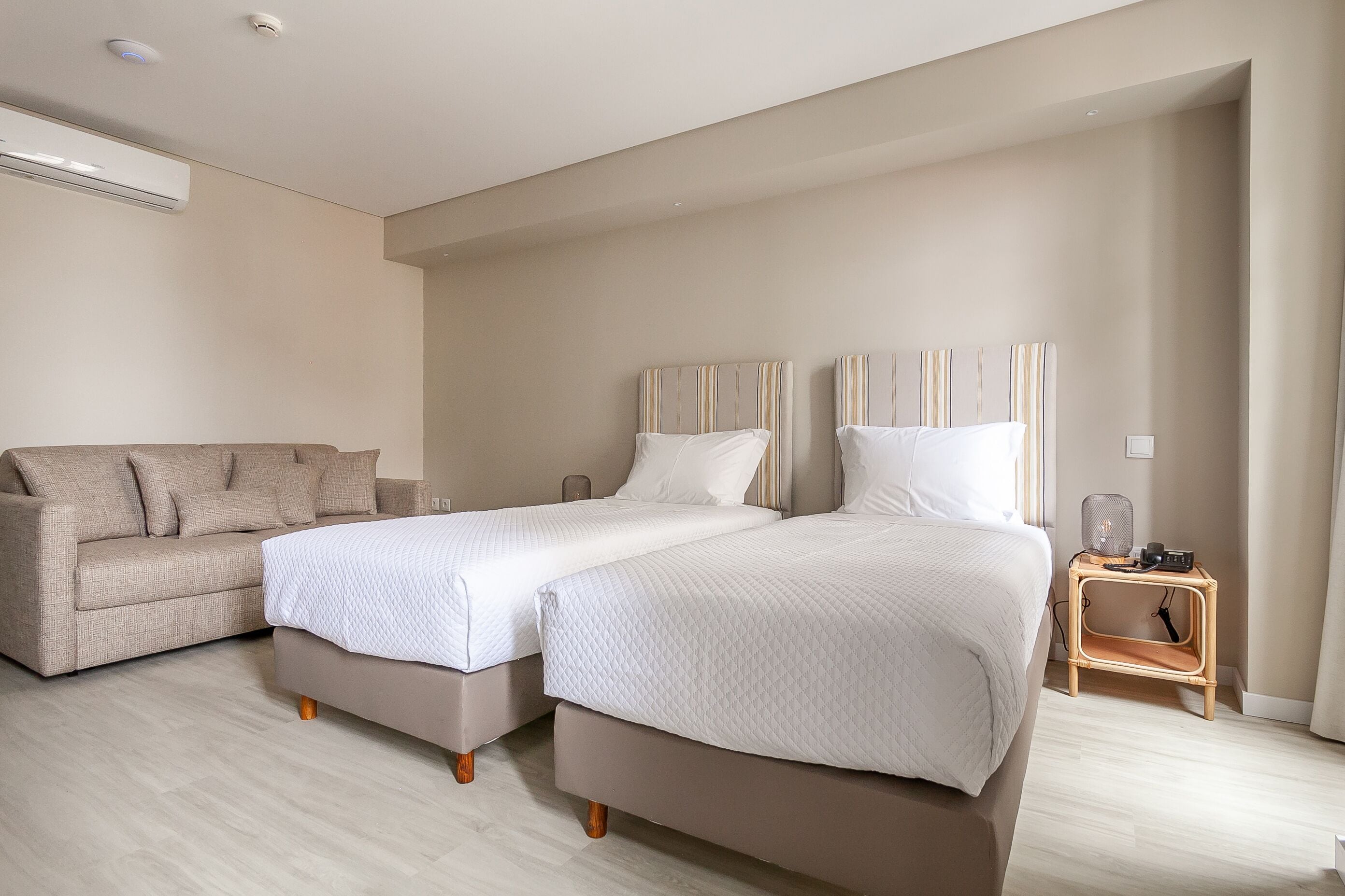 Hotellihuone, jossa on kaksi erillistä sänkyä, sohva, pieni pöytä ja ilmastointilaite.