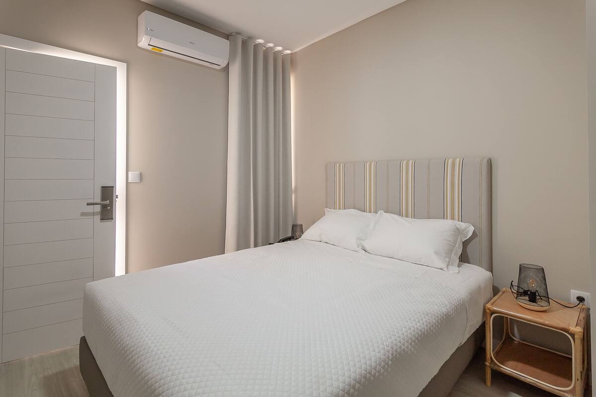 Una cama bien hecha con una colcha blanca acolchada, una mesita de noche con lámpara y un equipo de aire acondicionado en la pared.