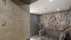 Interior moderno com parede de madeira exibindo nomes de quartos e indicações, um sofá cinza e um papel de parede estampado.