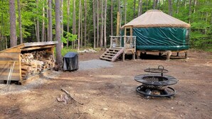 Un terrain de camping rustique doté d’un chalet en bois, d’un foyer et d’un espace de repos couvert, le tout entouré de grands arbres.